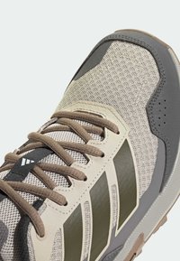 Κοντινό πλάνο μπεζ και γκρι αθλητικού παπουτσιού με καφέ κορδόνια και τρεις ελιάς πράσινες ρίγες στο πλάι, που δείχνει πλέγμα και συνθετικά υλικά.