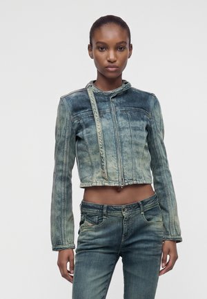 Donna che indossa una giacca di jeans corta sbiadita con cerniera e jeans blu sbiaditi abbinati, in piedi davanti a uno sfondo chiaro e uniforme.
