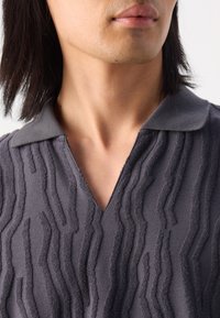 Maglione grigio con un motivo a onda testurizzato, caratterizzato da un design a V e un colletto morbido. Il tessuto appare peloso e tattile.
