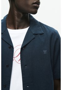 Cămașă cu mânecă scurtă de culoare bleumarin, dintr-un material texturat, având căptușeală de tip camp, nasturi și un tricou alb dedesubt, cu un imprimeu grafic.
