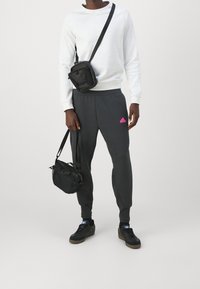 Grijze sweatpants met roze logo, witte sweatshirt en zwarte schoudertassen. Zwarte sneakers maken de outfit compleet. Gladde stof en ontspannen pasvorm.