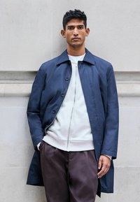 Palton ușor, de culoare bleu marin, cu închidere cu nasturi, guler mare și croială dreaptă; purtat peste un hanorac gri cu fermoar și pantaloni închiși la culoare.