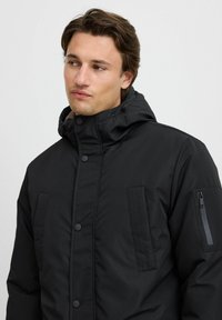 Sort pufferjakke med høj krave, knaplukning, forlommeskaber og lynlåslomme på ærmet. Glat tekstur og polstret design.