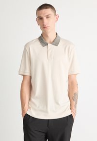 Beige polo skjorte med et mønstret grått krage, korte ermer og rett snitt. Har tre knapper foran og en glatt stofftekstur.