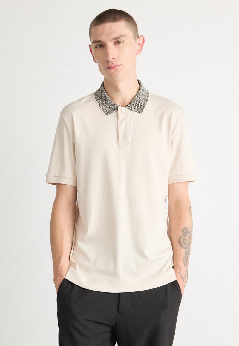 Beige polo skjorte med et mønstret grått krage, korte ermer og rett snitt. Har tre knapper foran og en glatt stofftekstur.