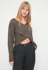 Brun ribbet strikket kort genser med V-hals og asymmetrisk nedre kant, kombinert med mørkegrå skreddersydde bukser med knappdetalj.