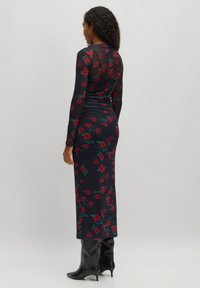 Langärmliges, tailliertes Kleid aus durchscheinendem schwarzem Material mit einem dunklen Blumenmuster in Rot und Grün. Knielang, kombiniert mit schwarzen Stiefeletten mit Absatz.