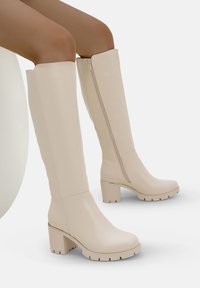 Botas beige hasta la rodilla con un acabado de cuero suave, tacones de bloque gruesos, cremalleras laterales y suelas de goma con relieve para mayor tracción.