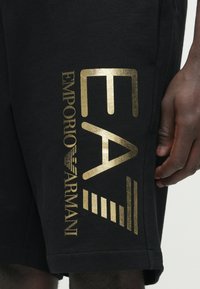 Fekete rövidnadrág fémes arany "EA" logóval és "Emporio Armani" felirattal, oldalán vízszintes csíkos mintával. Pamut anyag, laza szabás.