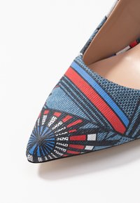 Chaussure à talon colorée avec un bout pointu, en tissu texturé bleu, et un motif géométrique avec des accents rouges, bleus et noirs.