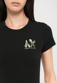 Juodas trumpomis rankovėmis marškinėliai su blizgiu auksiniu "A|X Armani Exchange" logotipu kairėje krūtinės pusėje. Lygus paviršius, prigludusi forma.