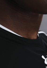 Κοντινό πλάνο του λαιμού και της γραμμής της γνάθου ενός ατόμου, που φοράει μαύρο πουκάμισο με λευκό λογότυπο Under Armour ορατό κοντά στον γιακά.