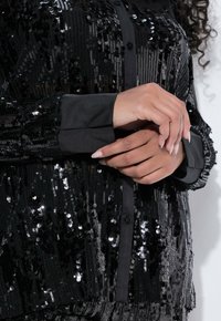 Black sequin shirt featuring a button-down front and contrasting black cuffs. Smooth texture with shiny sequins for a sparkling appearance.