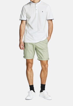 Kurzärmliges weißes Poloshirt mit schwarzen Akzenten, kombiniert mit hochgerollten hellgrünen Shorts und weißen Sneakern, was ein lässiges Outfit ergibt.