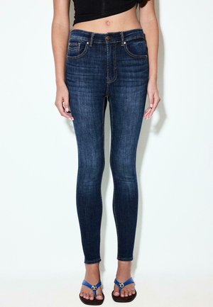 Femme portant un jean skinny bleu foncé, un crop top noir et des sandales plates bleues, debout devant un fond clair uni.
