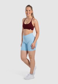 Rood sportbeha met dunne bandjes, gecombineerd met lichtblauwe shorts met hoge taille. Model draagt witte sneakers en staat tegen een neutrale achtergrond.