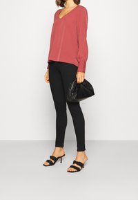 Eine bordeauxrot V-Ausschnitt-Bluse mit langen Ärmeln und Knopfdetails, kombiniert mit schwarzen Leggings und schwarzen Riemchen-Absatzsandalen.