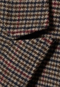 Puha gyapjú anyag, amelyen barna, tengerészkék, bézs és piros vonalakban houndstooth minta látható, hullámzó redőkkel és texturált szálakkal.