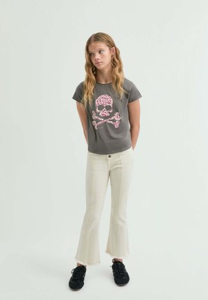 Mujer joven con cabello largo y ondulado que lleva una camiseta gris oscuro con una calavera floral rosa y huesos cruzados, vaqueros crema acampanados y zapatillas negras.