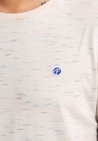 Lichtbeige t-shirt met een zachte textuur, voorzien van kleurrijke horizontale strepen en een klein blauw cirkelvormig logo op de borst.