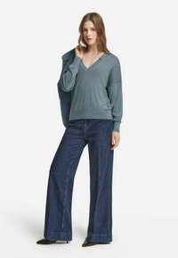 Ung kvinna med långt hår som bär en lös tealfärgad V-ringad tröja, vida mörkblå jeans och svarta spetsiga skor, håller en jacka över axeln.