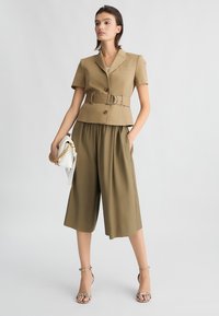 Blazer khaki con cintura e maniche corte, abbinato a pantaloni culotte larghi. Accessoriato con una borsa bianca e scarpe con tacco.