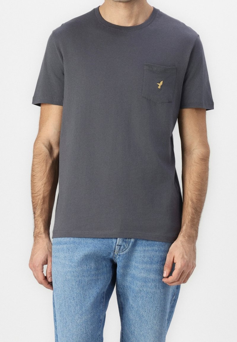 Homme portant un t-shirt à manches courtes gris foncé uni avec un petit logo d'oiseau jaune sur la poche poitrine gauche et un jean bleu clair.