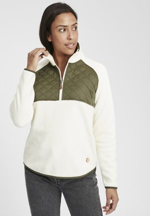 Fleece pullover in cream with a quilted olive green yoke, half-zip, and decorative logo button at the hem. Features contrasting trim.