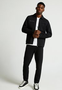 Zwarte zip-up jas met twee voortassen, gecombineerd met zwarte broek en zwart-witte sneakers. Effen wit t-shirt eronder.