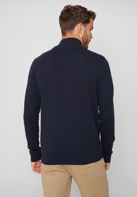 Dunkelblauer Strickpullover mit hohem Kragen, langen Ärmeln und gerippten Bündchen. Glatte Textur und taillierte Passform. Getragen mit hellbeigen Hosen.