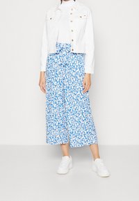 Witte spijkerjack met gouden knopen over een lange blauw-wit gebloemde rok met een strikceintuur, gecombineerd met witte sneakers.