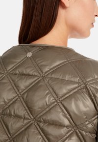 Gestepptes, olivgrünes Jackett mit Rautenmotiv, glänzender Oberfläche und einem runden Metalllogo-Akzent hinten in der Nähe des Kragens.