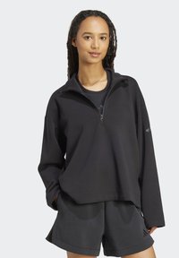 Crna zip-up hoodie izrađena od mekog i rastezljivog materijala s visokom kragnom; kombinira se s crnim kratkim hlačama koje imaju bočne džepove i akcentu s logom.