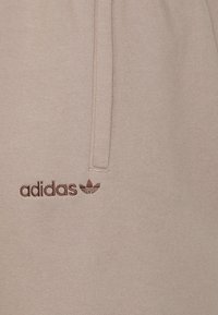 Beige mikina s měkkou texturou, na levé straně s hnědým logem "adidas" a akcentem ve tvaru trojlístku. Klasický střih a minimalistický design.
