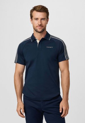 Homme portant un polo bleu marine avec des rayures blanches sur les épaules et le logo "Hackett", se tenant devant un fond gris clair uni.