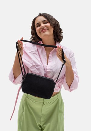Black suede crossbody bag with pink cord accent, rectangular shape, zipper closure, and adjustable strap, held by a person.