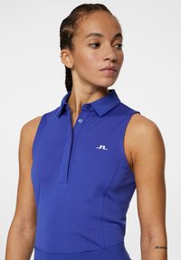 Lila ärmlös polo-t-shirt med krage, med en knappslå och en liten vit logotyp på vänster bröst. Slät tygtextur.