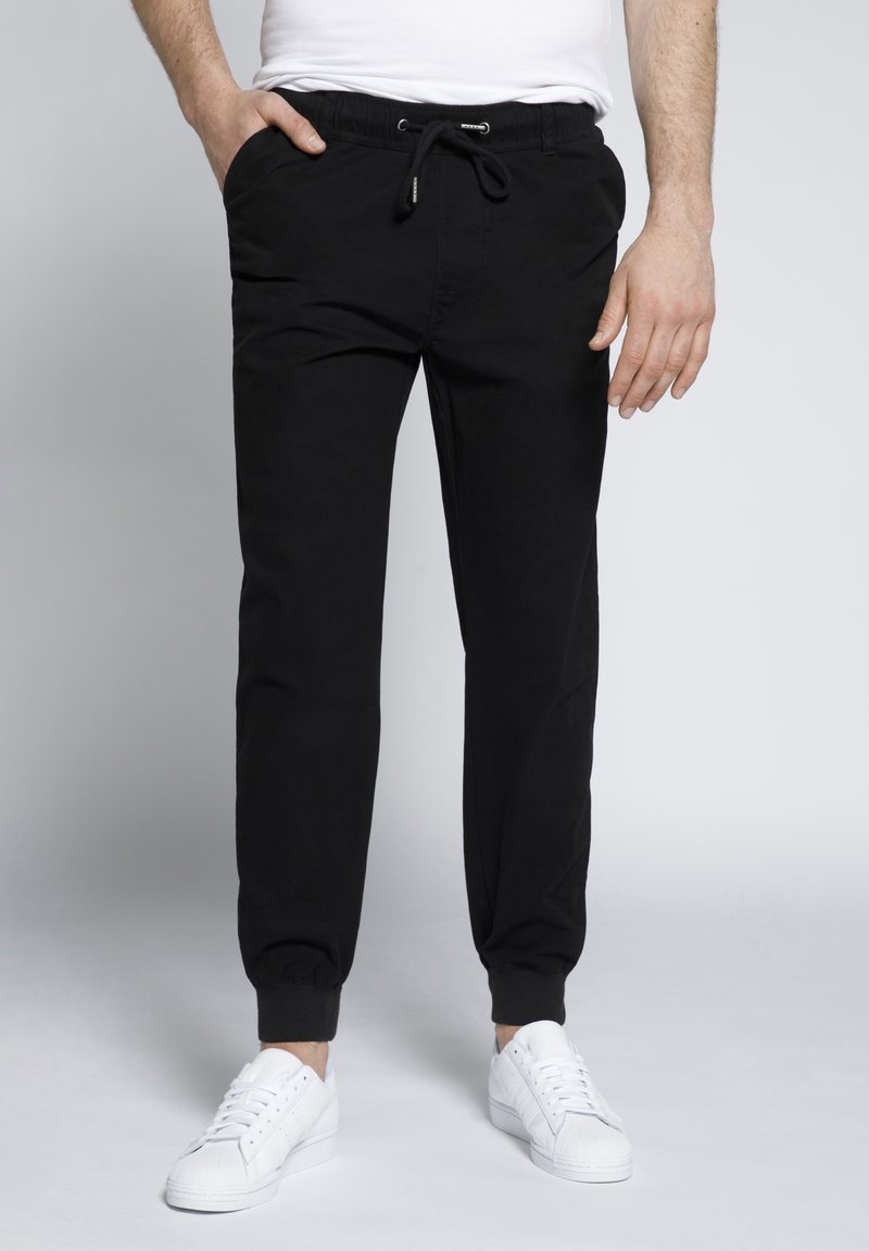 Pantaloni da jogging neri in cotone con vita elasticizzata, cordoncino, tasche laterali e polsini a costine; abbinati a sneakers bianche.