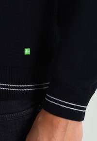 Námořnický svetr s bílými pruhy na manžetě, zelená nášivka s logem BOSS Hugo Boss, a část ruky v tmavých džínách.