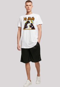 White cotton t-shirt featuring a colorful Def Leppard graphic in a triangular design, paired with black shorts and white sneakers.