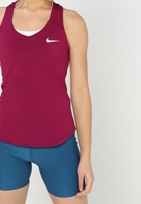 Top senza maniche Nike in bordeaux, con logo bianco, abbinato a pantaloni corti blu. Il tessuto appare liscio e con un design sportivo.