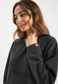 Zwarte sweatshirt met een ronde hals, van textuurstof, voorzien van een klein geborduurd logo en een decoratief rood label op de mouw.