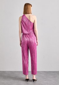 Combinaison fuchsia avec une texture brillante et plissée ; design asymétrique à une épaule ; haut court et pantalon droit ; portée avec des sandales à talons noires.