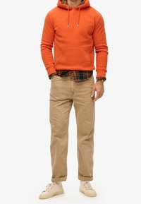 Sudadera con capucha naranja con bolsillo frontal, superpuesta sobre una camisa de franela a cuadros. Pantalones beige y zapatillas de color claro completan el conjunto.