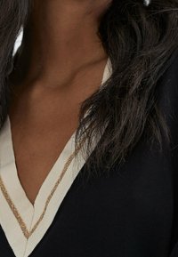Haut en tricot noir avec un décolleté en V profond orné d'une bordure blanche et d'une bande dorée. Texture douce visible autour de l'encolure.