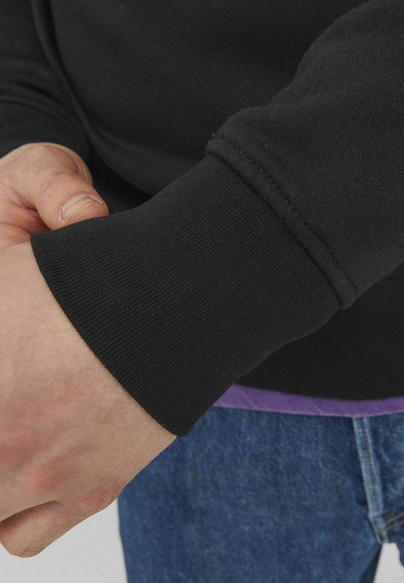 Zwarte sweatshirtmanchet met ribbeltextuur,close-upweergave; hand gepositioneerd nabij de manchet, spijkerbroek zichtbaar eronder.