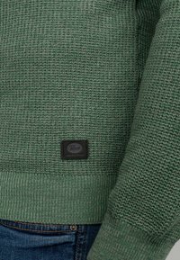 Maglione verde lavorato a maglia con un motivo testurizzato, orlo a coste e un'etichetta nera tessuta di lato. Abbinato a jeans blu.