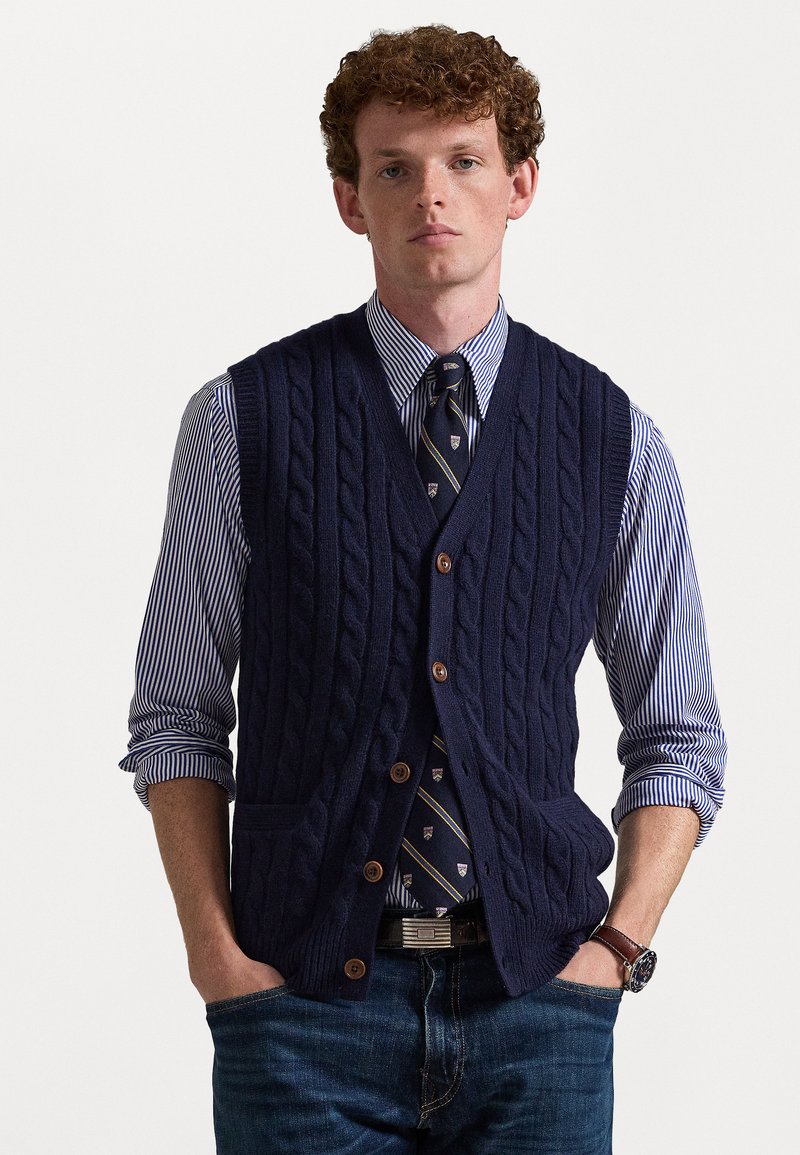 Marineblå strikket vest med kabelmønster og fem brune knapper. Bært over en stripete skjorte og et mønstrete slips, kombinert med jeans.