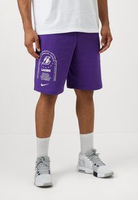 Lilla Nike shorts med Los Angeles Lakers branding, herunder tekst og logo. Båret med grå sneakers og hvide sokker.
