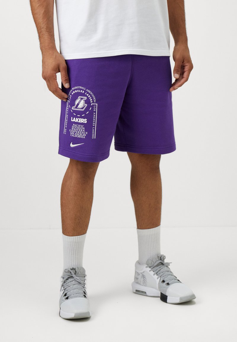 Calças curtas roxas da Nike apresentam a marca do Los Angeles Lakers, incluindo texto e logótipo. Usadas com ténis cinzentos e meias brancas.